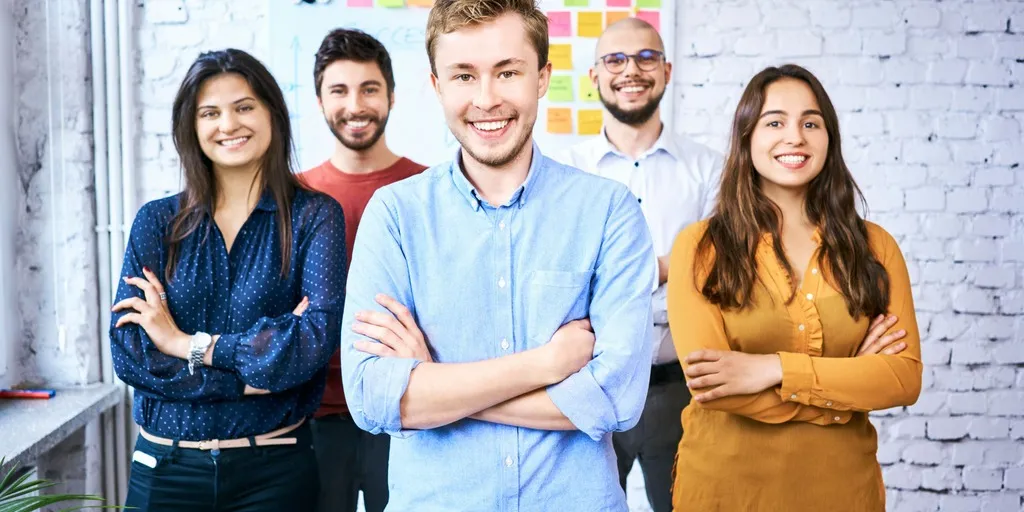 Eine Gruppe junger Menschen steht in einem Büro und schaut die Betrachtenden lachend an