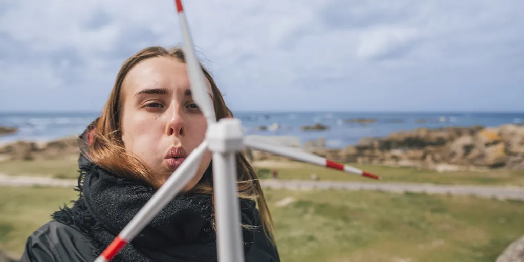 Eine Person steht im Freien, bläst in die Rotorblätter eines kleinen Windrades. Im Hintergrund sind das Meer und Felsen sichtbar. Die Person trägt einen schwarzen Schal und eine Jacke.