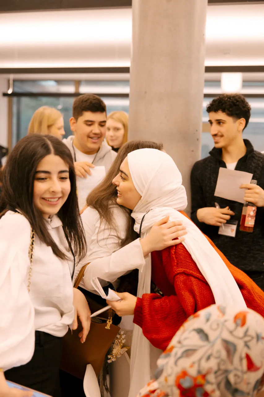 Junge Menschen begrüßen sich herzlich auf einem Event, eine Frau mit rotem Pullover und weißem Hijab umarmt eine andere Person