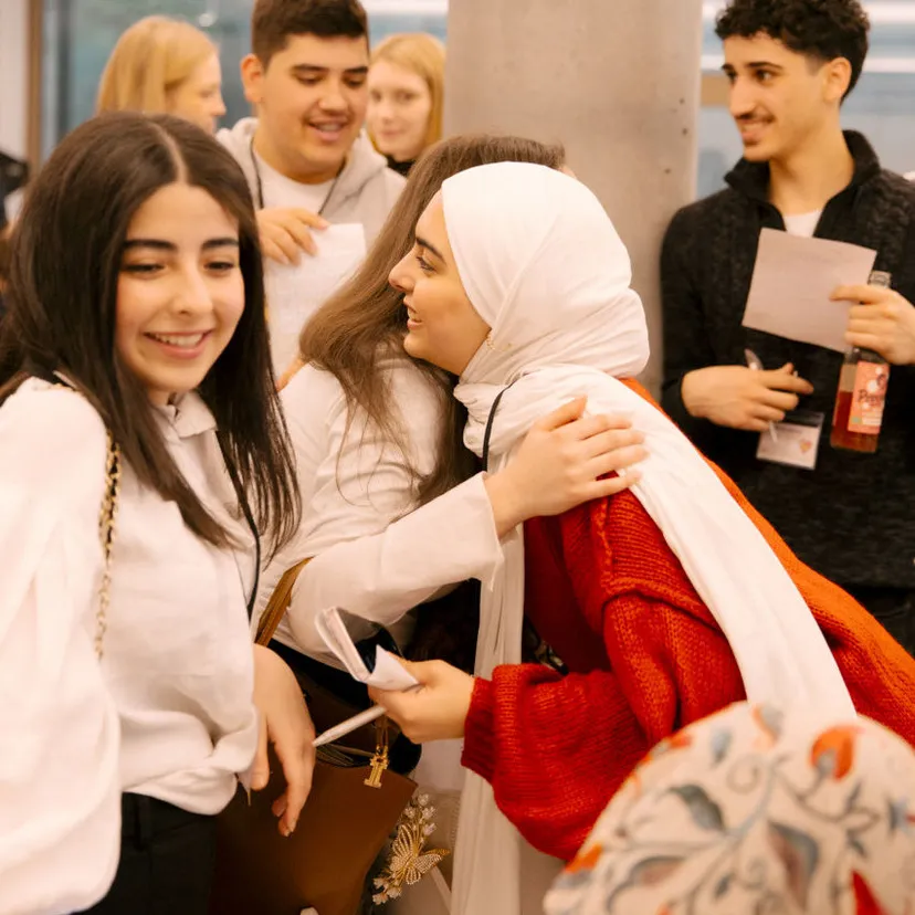 Junge Menschen begrüßen sich herzlich auf einem Event, eine Frau mit rotem Pullover und weißem Hijab umarmt eine andere Person