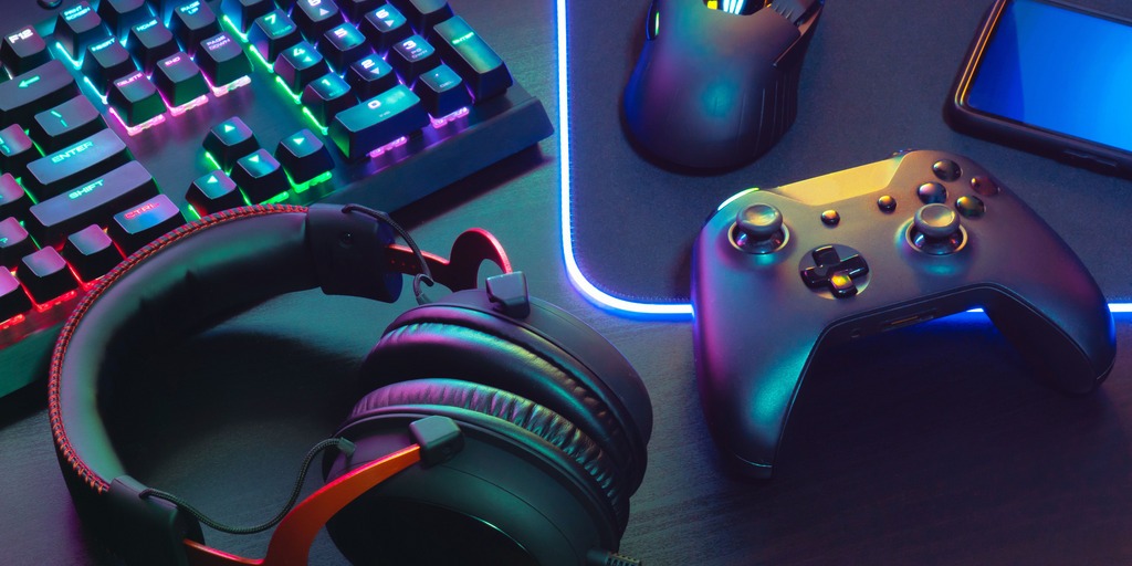 Das Bild zeigt ein Set aus Gaming-Zubehör auf einem Schreibtisch: eine beleuchtete mechanische Tastatur, eine Gaming-Maus auf einem leuchtenden Mauspad, ein schwarzer Gamecontroller, ein Smartphone und ein Paar Over-Ear-Kopfhörer. Alle Geräte sind in farbigem RGB-Licht inszeniert.