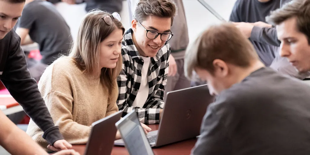 Mehrere junge Menschen sitzen um einen Tisch herum und blicken auf die Bildschirme der vor ihnen stehenden Laptops.